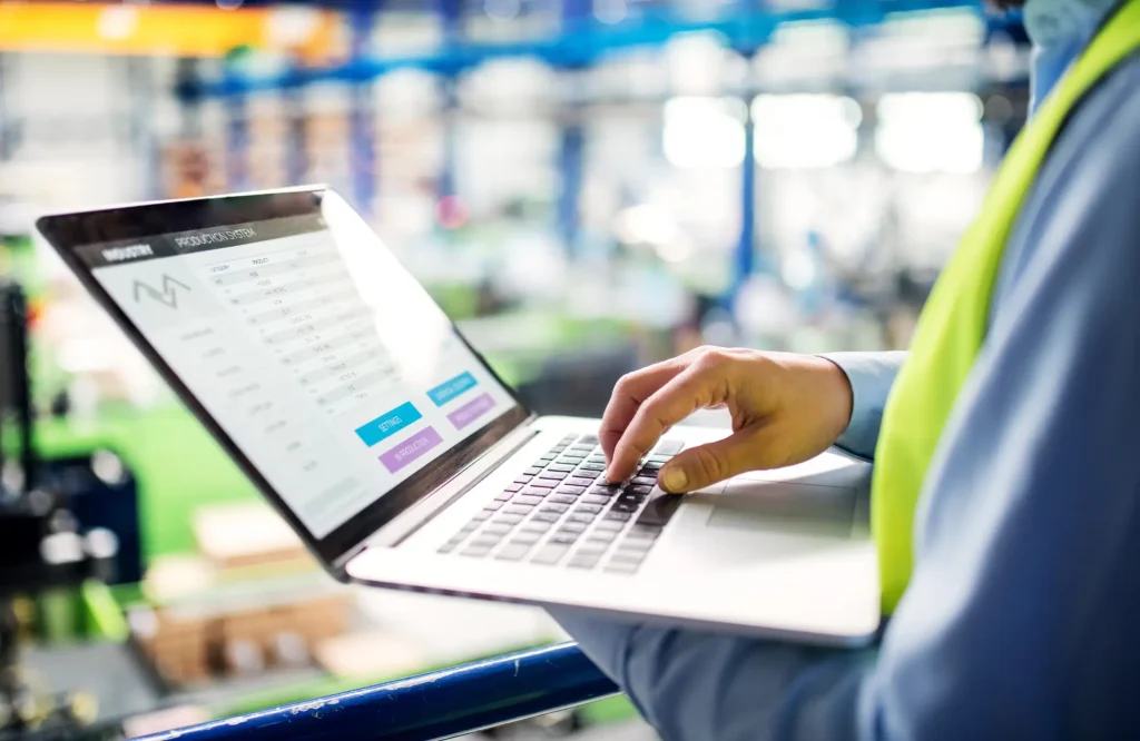 Tuotannonhallinnan ja raportoinnin digitalisoint -blogon kansikuva, jossa henkilö käyttää kannettavaa tietokonetta tehtaassa.